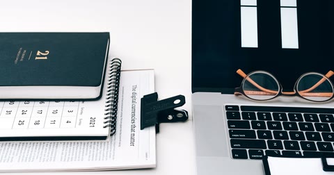 Agenda de capa preta sobre um calendário de mesa, ao lado de um notebook aberto com um par de óculos