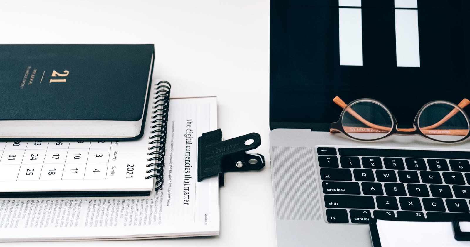 Agenda de capa preta sobre um calendário de mesa, ao lado de um notebook aberto com um par de óculos