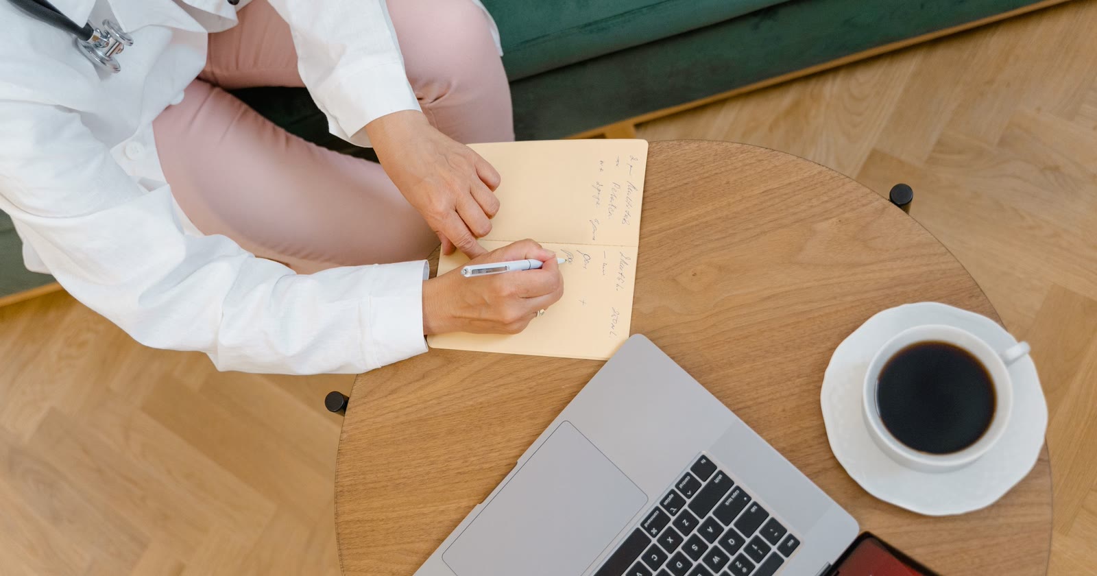 Profissional de jaleco branco anotando em um caderno ao lado de um notebook e de uma xícara de café