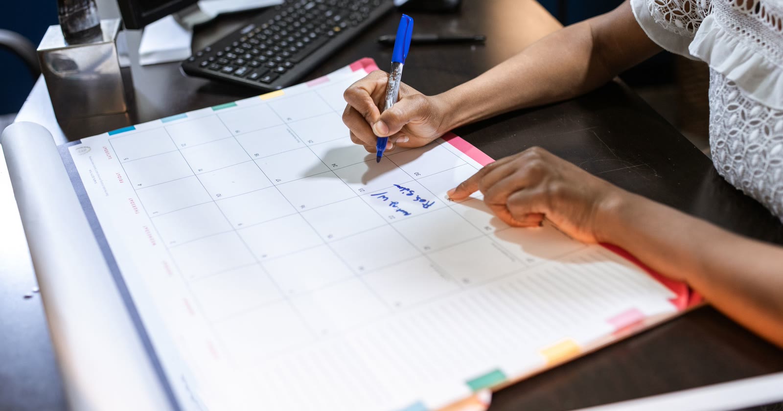 Mãos anotando um compromisso com caneta azul em um calendário de mesa, ao lado de um teclado