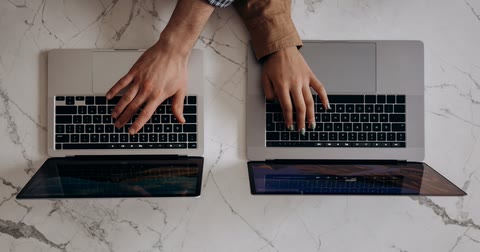 Duas mãos digitando em dois notebooks abertos lado a lado sobre uma mesa de mármore branco