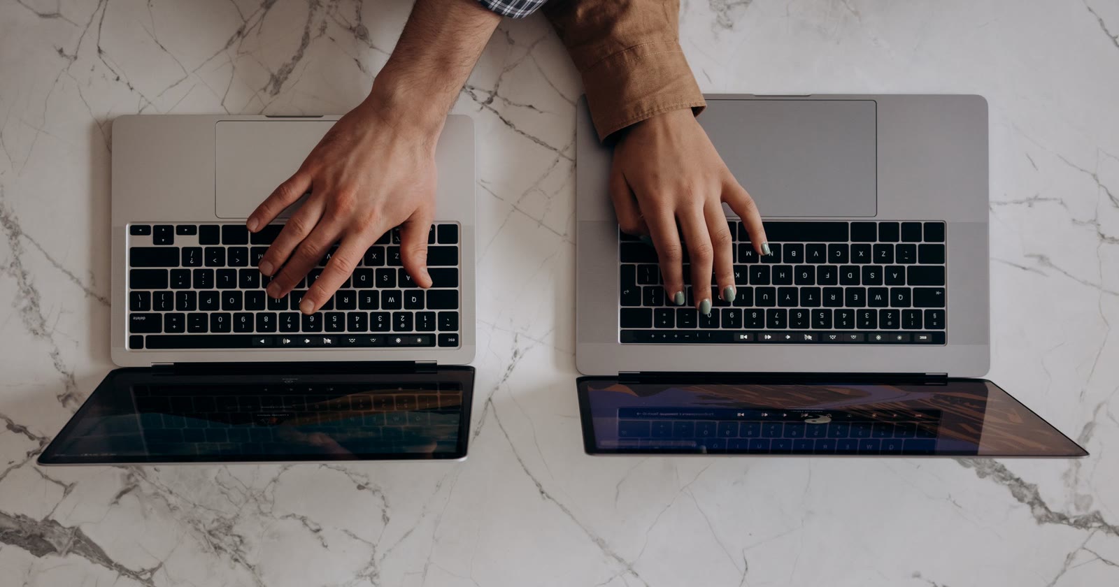 Duas mãos digitando em dois notebooks abertos lado a lado sobre uma mesa de mármore branco