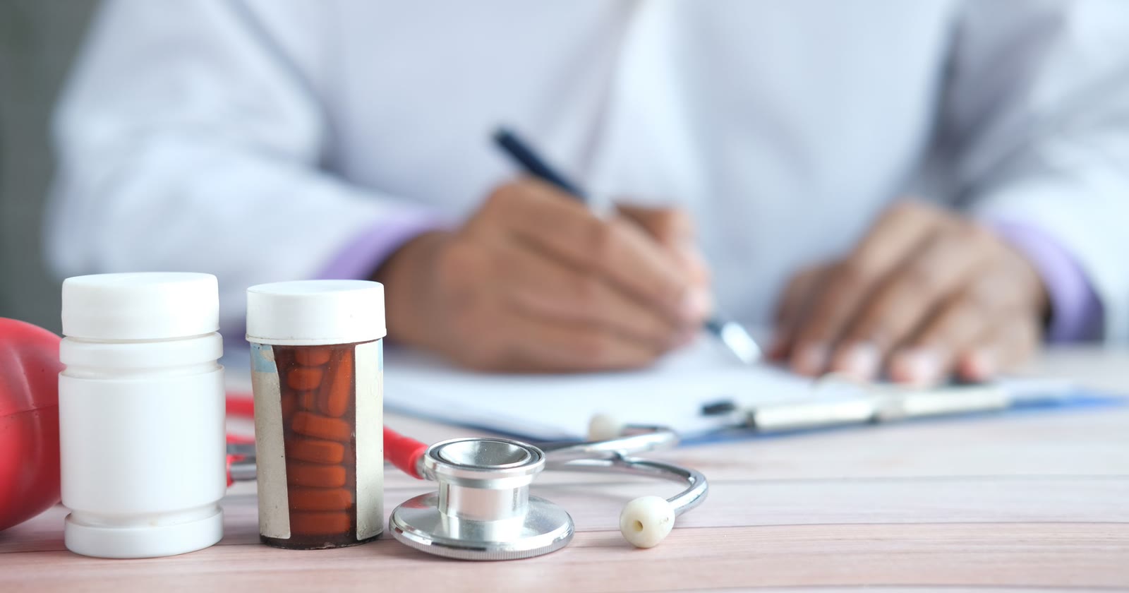 Mãos de um médico de jaleco escrevendo em uma prancheta, com estetoscópio e dois frascos de remédio sem rótulo sobre a mesa