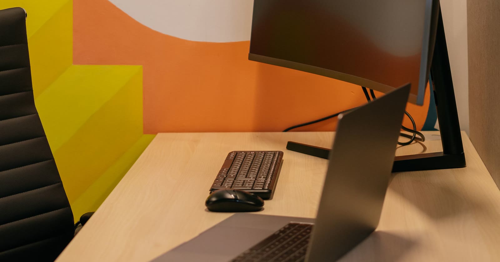 Mesa de trabalho com monitor, teclado e notebook aberto, sem ninguém na sala