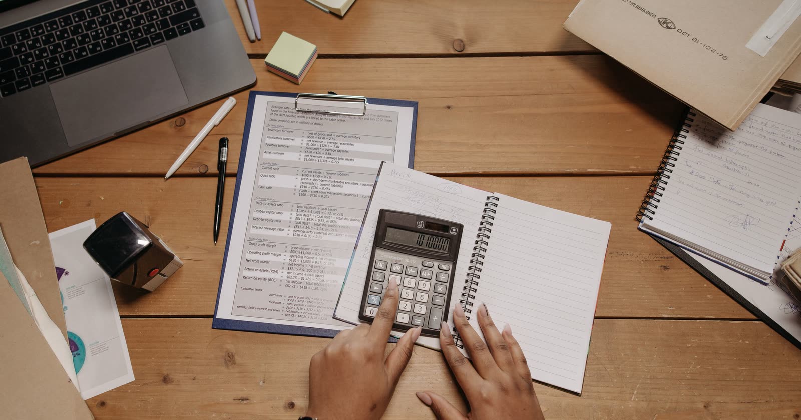 Mãos usando calculadora sobre mesa de madeira com prancheta, caderno, canetas e notebook