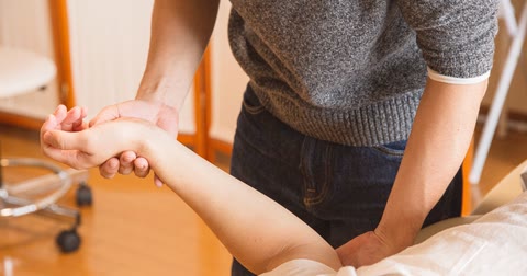 Mãos de um profissional apoiando o punho e o cotovelo de uma pessoa deitada durante um atendimento, sem rostos visíveis