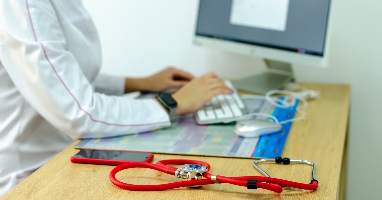 Profissional de jaleco digita no computador da clínica, com estetoscópio e celular sobre a mesa