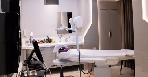 Sala de procedimentos de uma clínica de estética vazia, com maca, luminária e bancada com produtos